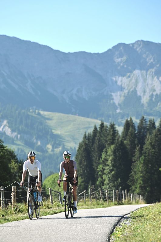 Ein Mann und eine Frau beim Rennrad fahren in Bad Hindelang auf einem asphaltierten Weg entlang eines Holzzauns.