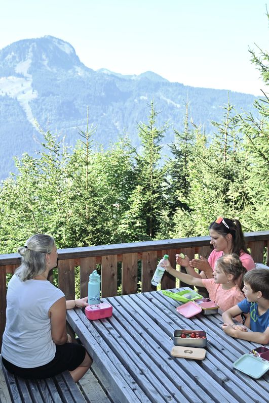 Eine Familie sitzt an einem Picknicktisch auf dem Geologischen Lehrpfad in Bad Hindelang und genießt die Mittagspause mit einem atemberaubenden Blick auf die majestätischen Berge und grünen Wälder.