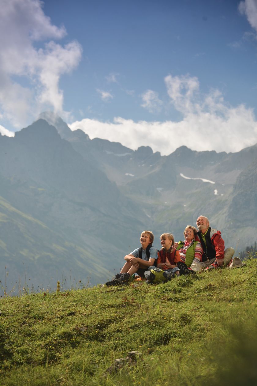 Ein älterer Mann und eine ältere Frau, ein Junge und ein Mädchen sitzen auf einer grünen Bergwiese und schauen in die Ferne, im Hintergrund sind Berge zu sehen.