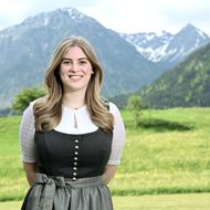 Eine lächelnde Mitarbeiterin der Tourist Information Bad Hindelang trägt eine traditionelle Tracht vor einer malerischen Berglandschaft mit grünen Wiesen und schneebedeckten Gipfeln.