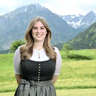 Eine lächelnde Mitarbeiterin der Tourist Information Bad Hindelang trägt eine traditionelle Tracht vor einer malerischen Berglandschaft mit grünen Wiesen und schneebedeckten Gipfeln.