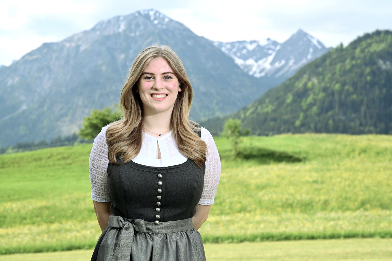 Eine lächelnde Mitarbeiterin der Tourist Information Bad Hindelang trägt eine traditionelle Tracht vor einer malerischen Berglandschaft mit grünen Wiesen und schneebedeckten Gipfeln.