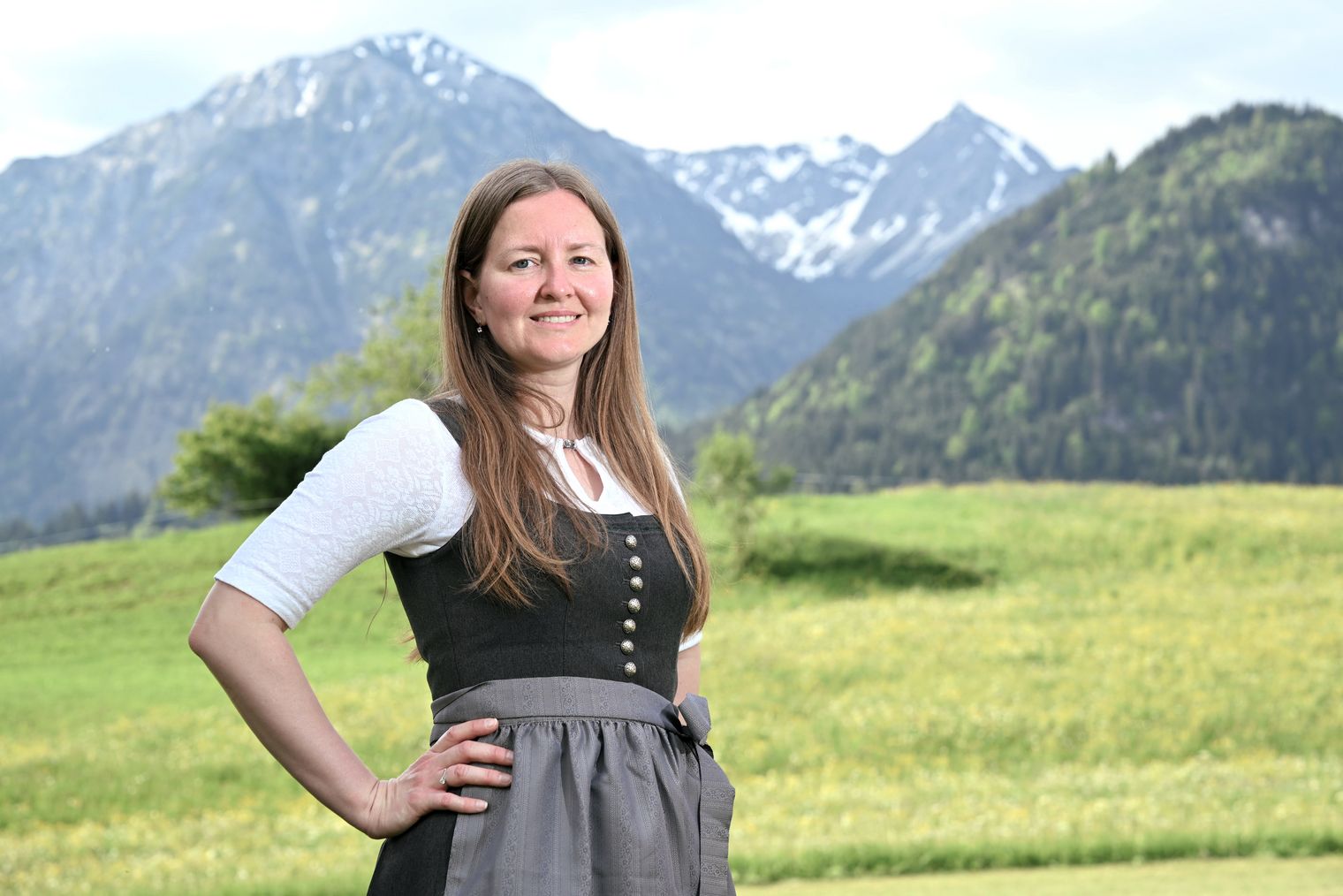 Eine lächelnde Mitarbeiterin der Tourist Information Bad Hindelang trägt eine traditionelle Tracht vor einer malerischen Berglandschaft mit grünen Wiesen und schneebedeckten Gipfeln.