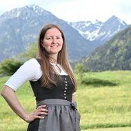 Eine lächelnde Mitarbeiterin der Tourist Information Bad Hindelang trägt eine traditionelle Tracht vor einer malerischen Berglandschaft mit grünen Wiesen und schneebedeckten Gipfeln.