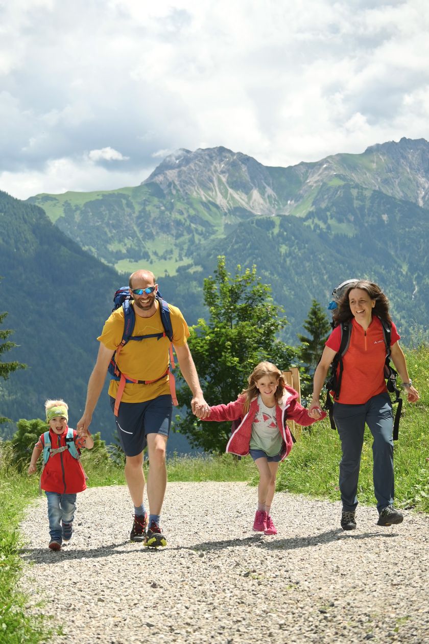Eine Familie in Wanderkleidung mit einem Mann, einer Frau, einem Mädchen und einem Jungen läuft Hand in Hand auf dem Burgschrofenweg in Bad Hindelang im Algäu.