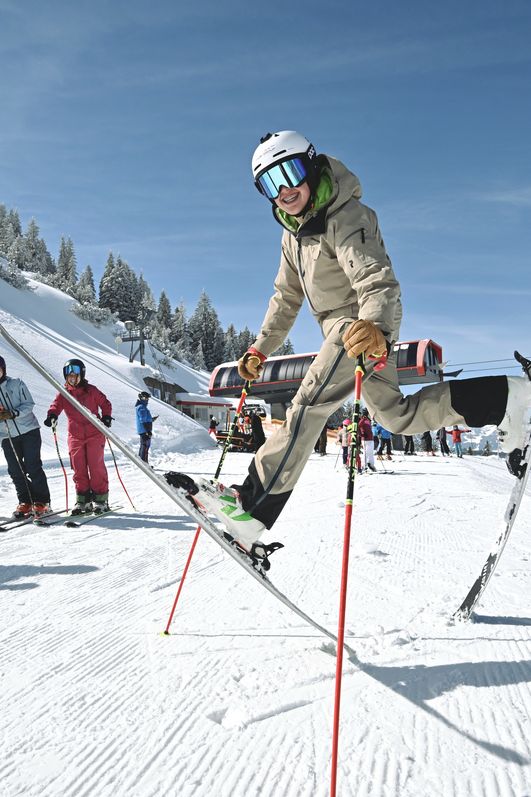Ein Skifahrer springt auf einem Ski im Vordergrund, während andere beim Skifahren am Oberjoch auf der Piste stehen und zuschauen.