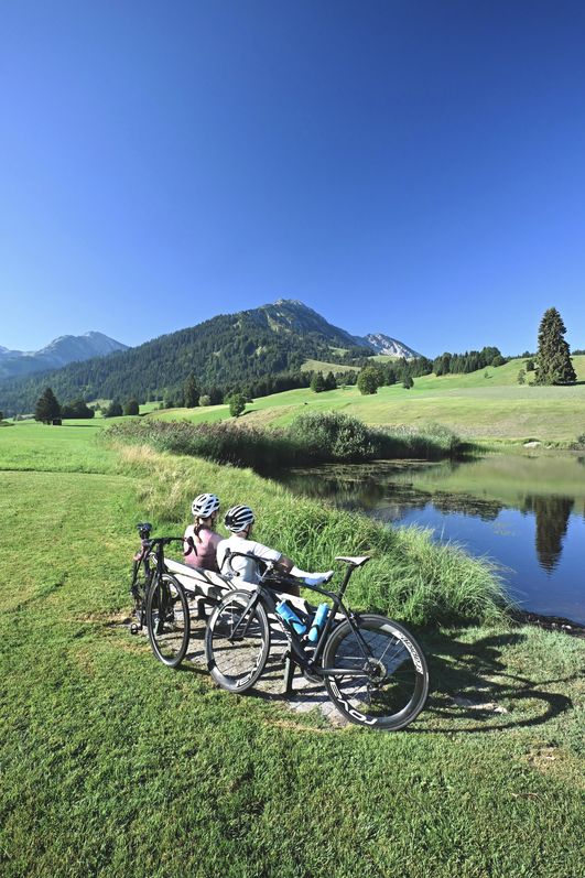 Erholung beim Rennrad fahren in Bad Hindelang: Zwei Sportler sitzen auf einer Bank an einem See im Grünen.