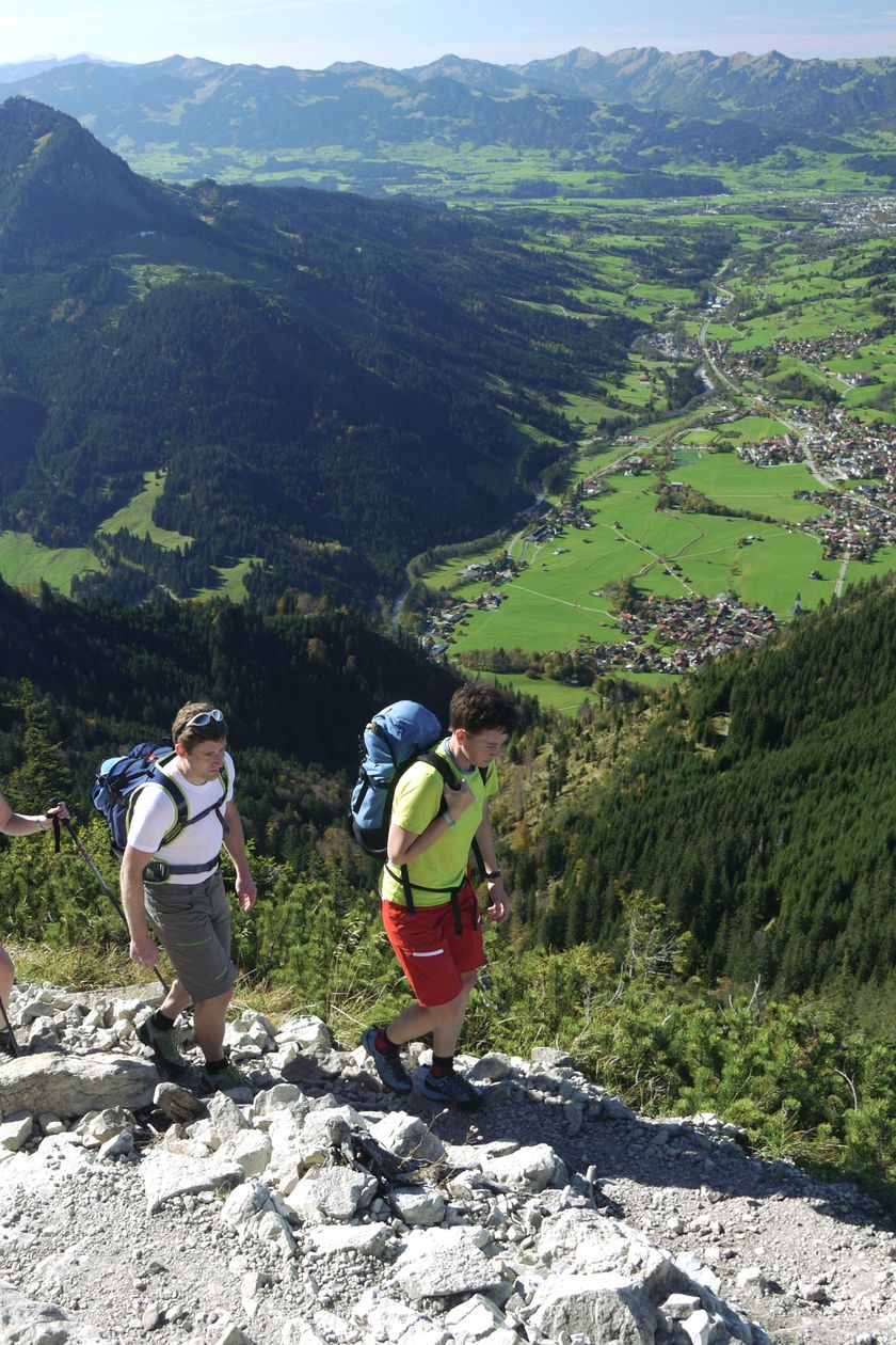 Drei Wanderer laufen auf einem steinigen Pfad am Iseler, im Hintergrund ist Bad Hindelang zu sehen.