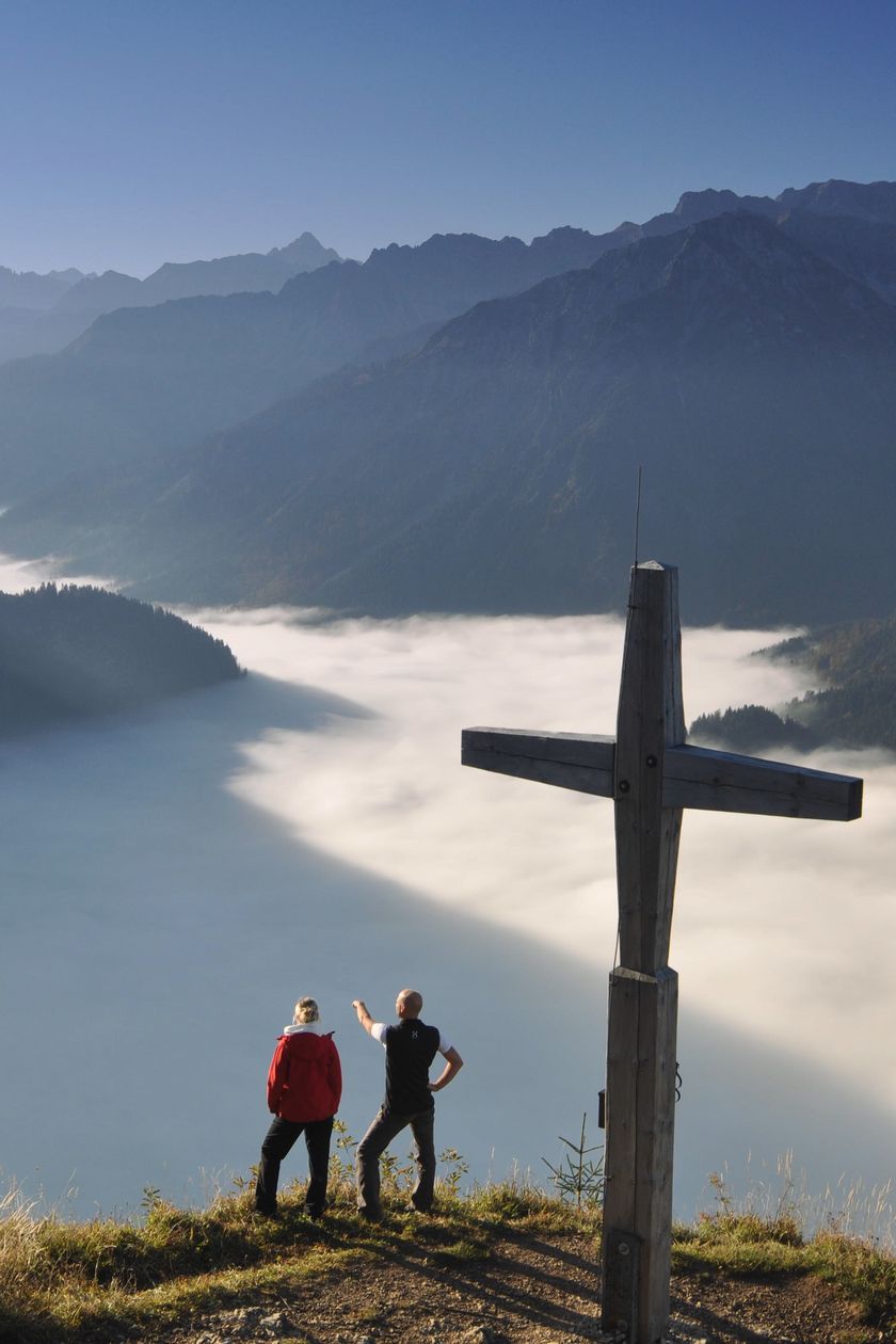 Zwei Wanderer stehen auf dem Hirschberg an dem Gipfelkreuz, vor ihnen ist ein Wolkenmeer zwischen den Bergen in Bad Hindelang im Allgäu zu sehen.