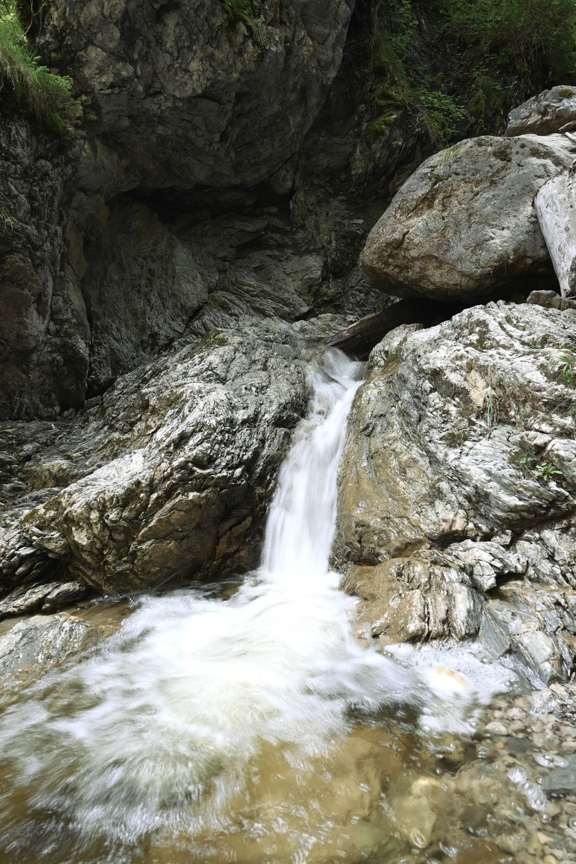 Das klare Wasser des Hirschbachs fließt über eine moosbewachsene Felsstufe, die eine kleine Kaskade bildet, während der Bach durch einen felsigen Einschnitt in Bad Hindelang weiterfließt.