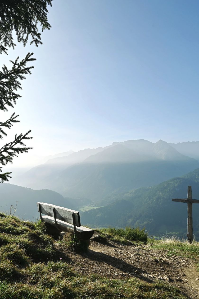 Malerischer Blick vom Gipfel des Hirschbergs in Bad Hindelang mit einer Bank und einem Holzkreuz im Sonnenlicht.