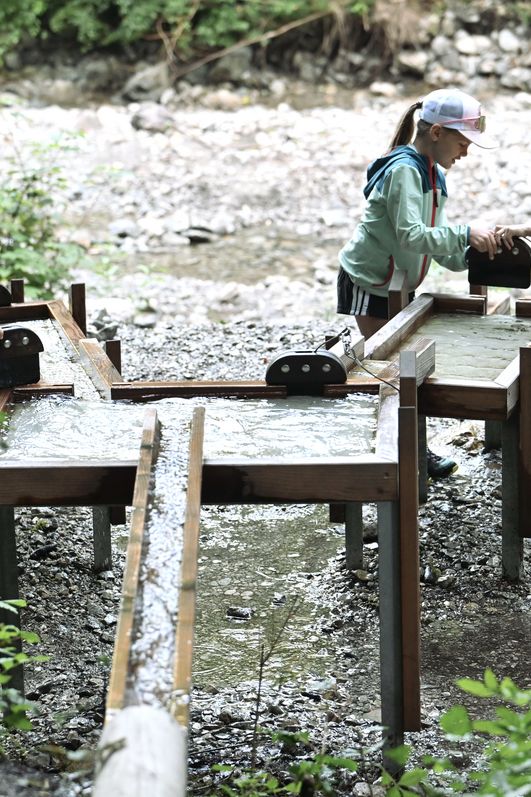 Drei Kinder spielen an einer hölzernen Wasseranlage mit Pumpen und Rinnen auf dem Geologischen Lehrpfad in Bad Hindelang, inmitten einer steinigen Bachlandschaft.