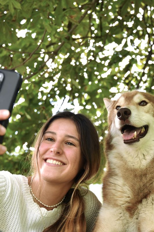 Eine junge Frau lächelt und macht ein Selfie mit einem braun-weißen Husky als Teil des Huskykuscheln-Erlebnisses in Bad Hindelang in der Natur.