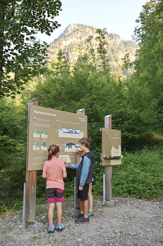 Zwei Kinder stehen vor einer Informationstafel zum Geologischen Lehrpfad in Bad Hindelang und studieren die Plattenbewegung, umgeben von Bäumen und Bergen.