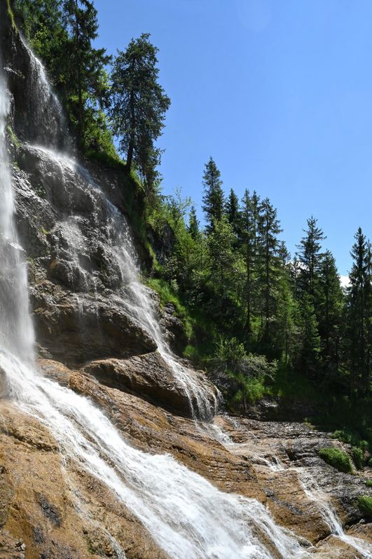 Ein Wasserfall rauscht über Felsen herab umringt von Wäldern, im Hintergrund ist ein Berg und blauer Himmel zu sehen.