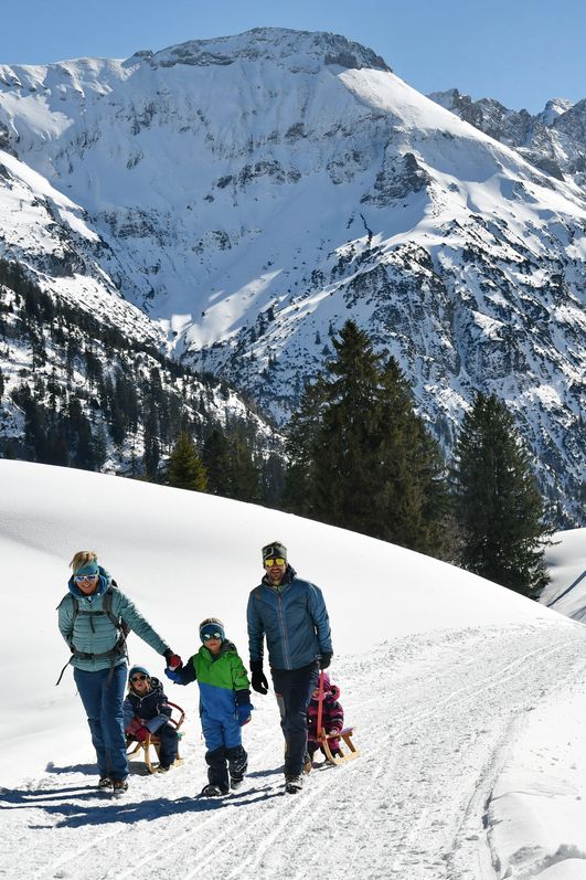 Eine Familie mit Mutter, Vater und drei Kindern und zwei Schlitten läuft auf einem präparierten Weg inmitten einer Weißen Winterlandschaft, im Hintergrund sind schneebedeckte Berge zu sehen.