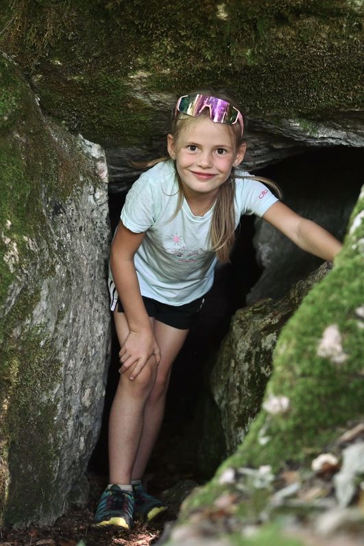Ein junges Mädchen in Freizeitkleidung steht am Geologischen Lehrpfad in Bad Hindelang in einem moosbewachsenen Felsdurchgang und lächelt in die Kamera.